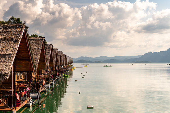 Serene row of stilted bungalows with thatched roofs on a tranquil lake at sunset, kayaks dot the calm water with distant misty mountains under a dramatic cloudy sky.