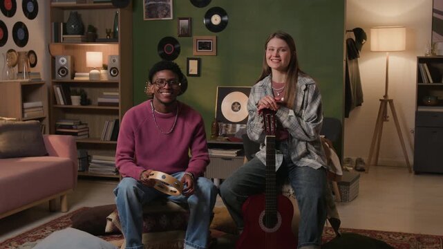 Wide angle shot of young adult female guitarist smiling at camera posing together with Black male tambourine player during home rehearsal session, slow motion