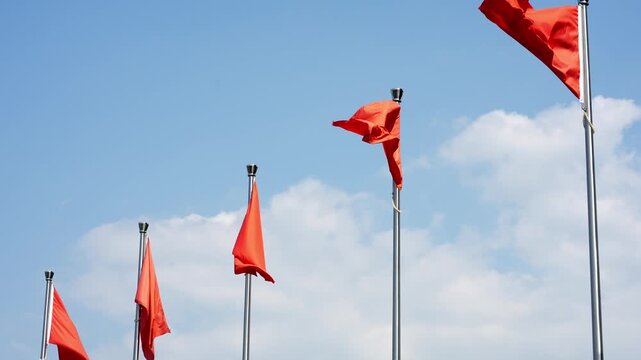 Five red flags waving in the wind against a blue sky with white clouds