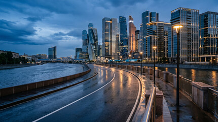 Obraz premium Sleek urban riverside road curving past modern high rise skyscrapers with wet asphalt street reflections