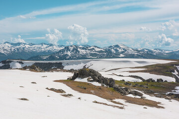 Scenic alpine landscape with sunlit snowy field on stony hill with view to big mountain range with forest and snow far away under cloudy sky. Stone outcrops among snows in sunlight in high mountains. © Daniil
