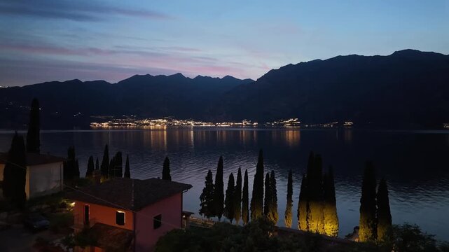 Evening dusk view over Garda lake in Malcesine with illuminated houses, reflected lights on water, serene mountains, travel tourism scenery