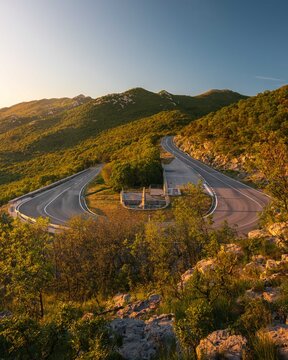 Aerial view of a winding mountain road with a sharp hairpin turn and a stone monument surrounded by lush green hills during golden hour in Rijeka, Primorje-Gorski Kotar, Croatia.
