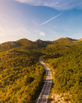 Aerial view of a winding road through lush green forested hills under a clear blue sky with a vapor trail during golden hour Rijeka, Primorje-Gorski Kotar, Croatia.