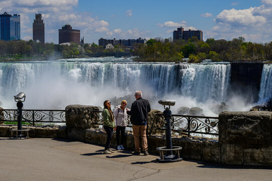 Two tourists with guide viewing Niagara Falls from Canadian side observation area, spring, with American skyline visible in background.