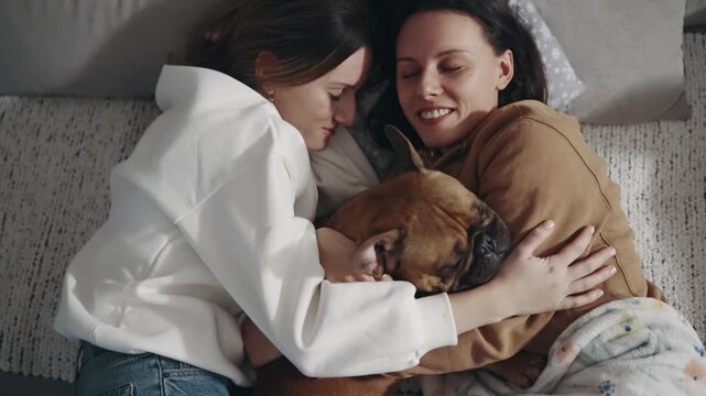 Two women are hugging a French bulldog dog while laying on a bed. Scene is warm and affectionate