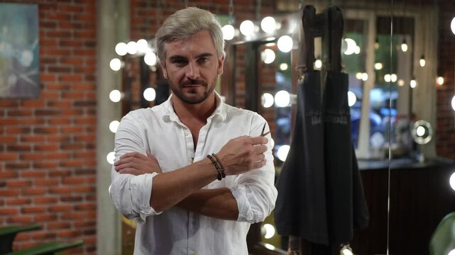 Portrait of a confident mid-adult male hairdresser with silver hair standing in his modern salon with his arms crossed, smiling