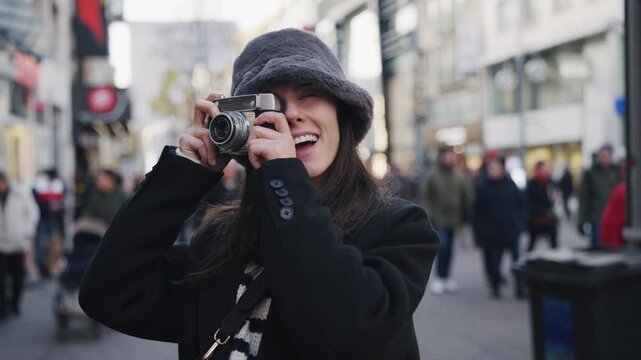 Happy smiling woman wearing a gray hat use vintage old camera taking pictures from at Christmas market in Germany. She is surrounded by people on a street. Scene is cheerful and lighthearted