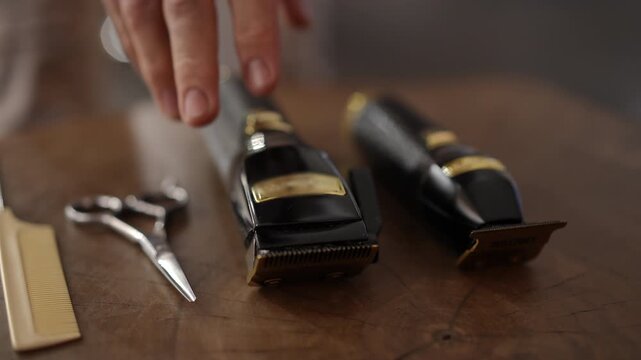 Professional male hairstylist picking up an electric hair clipper from a wooden table with other barber tools like a trimmer and scissors
