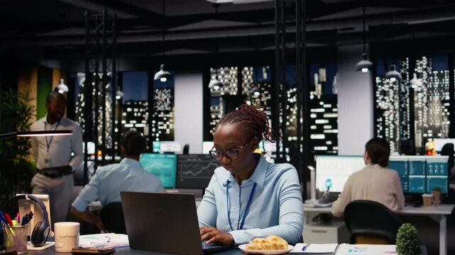 African American woman struggling with compulsive overeating, working on laptop. Employee with unhealthy eating habits serving junk food with carbs, reflecting mental health and burnout.