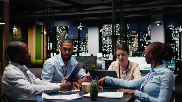 Diverse team of employees in office looking over paperwork documents late at night. Coworkers at work cooperating, analyzing company budget reports documentation files before deadline
