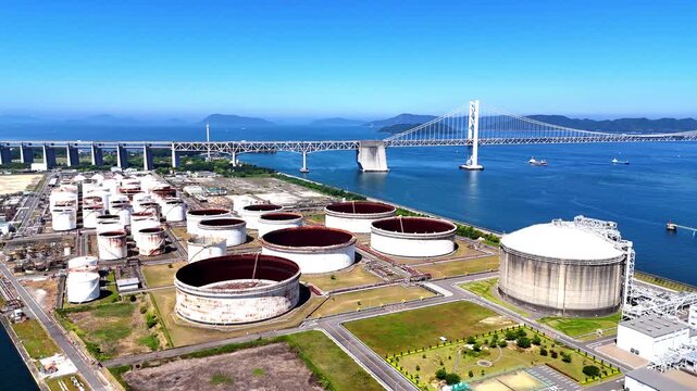 Crystal clear 4K aerial establishing shot of a modern coastal tank farm in Japan, highlighting large LNG and oil storage facilities set against the calm blue ocean and a suspension bridge.
