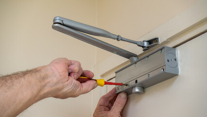 Building maintenance engineer adjusting an automatic door closer on a fire door using a screwdriver. Fire door compliance in commercial buildings. © RCH Photographic