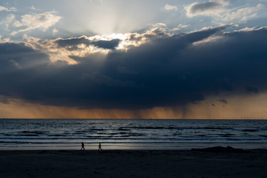 View of Strand Wijk aan Zee with silhouettes of people walking along the shore under dramatic sun rays breaking through dark clouds Wijk aan Zee, North Holland, Netherlands.