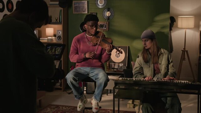 Backstage shot of videographer filming live performance of two young adult musicians playing synth and violin in home studio setup