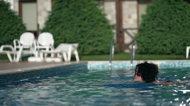A woman is swimming in a pool at sunset enjoying the summer vacation. She is wearing sunglasses and a black swimsuit. The pool is surrounded by trees and chairs