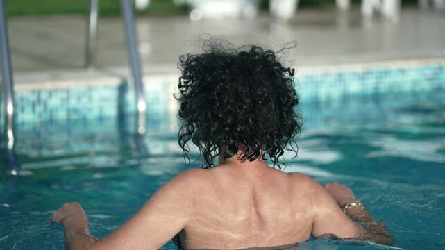 Hispanic woman walks on metal ladder stairs in swimming pool, wearing only a bikini top and no shirt, slow motion