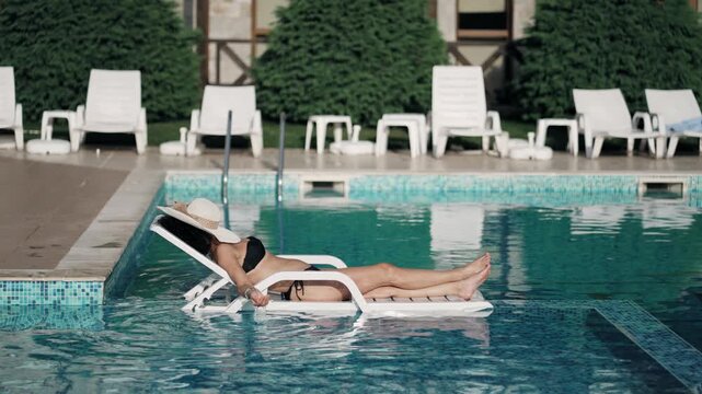Fit woman is laying in a pool chair, wearing a black bikini and a white hat over her face. The scene is relaxing and peaceful, with the woman enjoying her time in the water