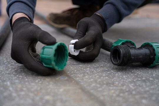 Hands in black work gloves holding a green locking nut and a white plastic compression ring next to a disassembled tee fitting during plumbing pipeline installation.