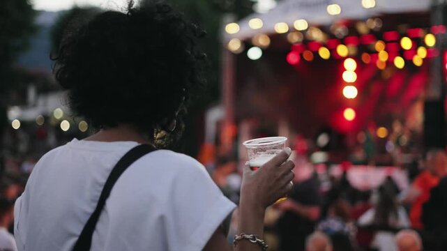 A woman is holding a cup of beer while standing at concert. Concept of enjoyment and relaxation, as the woman is likely at a social event or festival. The crowd around her suggests a lively atmosphere