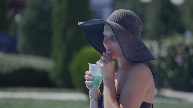 A woman in a black bikini is sitting in the sun with a cup of green liquid mojito cocktail in her hand