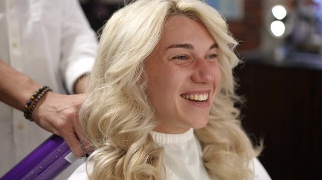 Professional hairstylist applying spray to a young happy woman's blonde curly hairstyle. Finishing touches for a beautiful coiffure in a salon