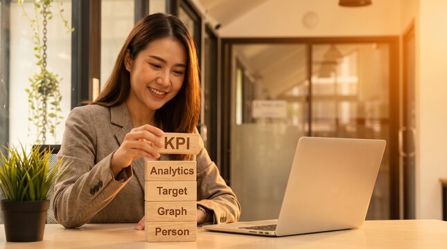 Businesswoman presents a KPI block stack beside a laptop in a warm office during late afternoon light.