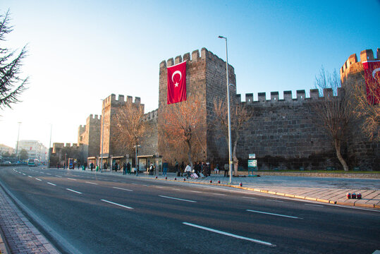 View of Kayseri Castle with stone walls and Turkish flags under a clear blue sky next to a city street with people in Kayseri, Kayseri, Turkiye.