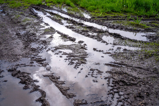 Muddy country road with deep water puddles and tire tracks after heavy rain next to green grass field.