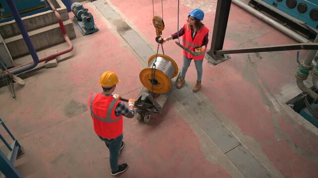 Two workers in hard hats and safety vests maneuvering a large spool of cable on a factory floor