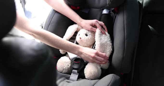 Danger prevention. Woman fastening toy bunny in child safety seat inside car, closeup