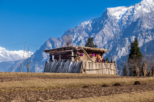 View of a traditional wooden house with people on the porch against the backdrop of snow-capped mountains and a clear blue sky in Arang Kel, Azad Kashmir, Pakistan.