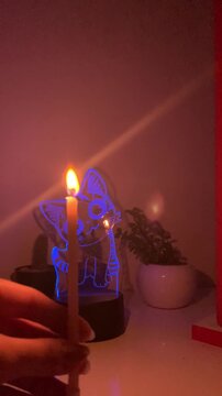 Person's hand lighting a white candle with a lighter in a dark room with a glowing blue neon cat lamp in the background, vertical 9:16 video.