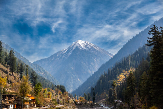 View of the snow-capped mountain peak of Neelum Valley surrounded by dense pine forests and a small village under a blue sky in Arang Kel, Azad Kashmir, Pakistan.