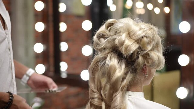 Professional male hairstylist creating an elaborate curly updo for a female client. Beauty salon interior with vanity mirror lights