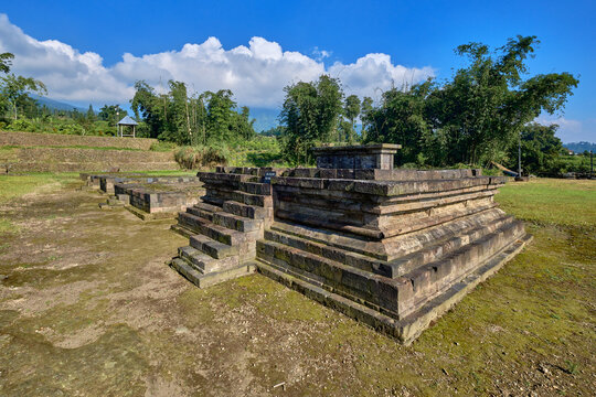 View of the ancient stone structures of Situs Liyangan archaeological site surrounded by lush greenery and trees under a blue sky in Temanggung, Central Java, Indonesia.