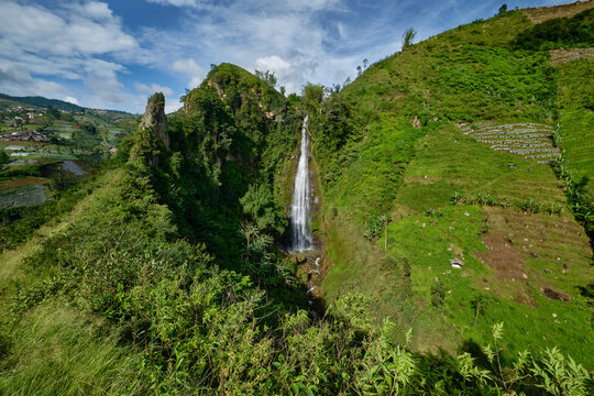 View of Curug Surodipo waterfall cascading down lush green cliffs surrounded by terraced agricultural fields under a blue sky in Temanggung, Central Java, Indonesia.