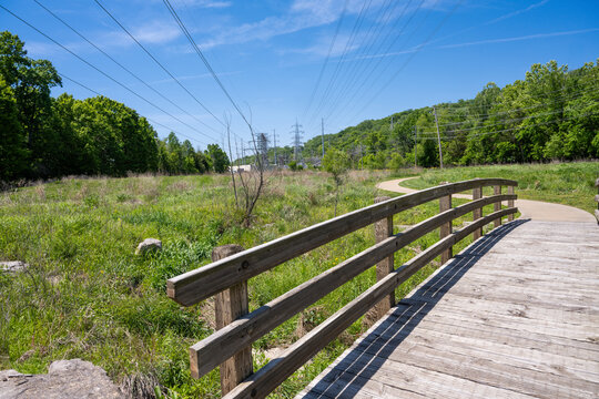 View of Greentree Park featuring a wooden bridge over a grassy field with power lines and lush trees under a clear blue sky Kirkwood, Missouri, United States.