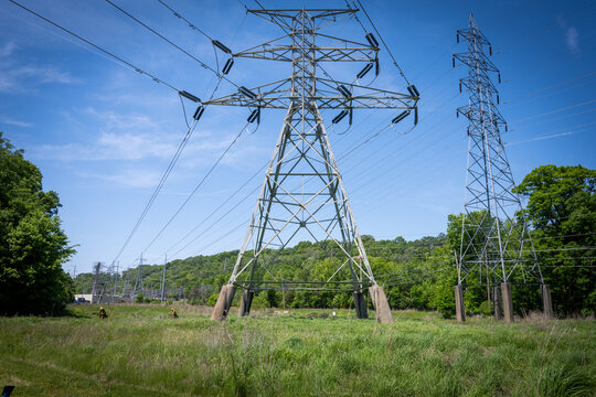 View of high-voltage power lines and electricity pylons at Greentree Park with workers in safety gear standing in a grassy field near trees in Kirkwood, Missouri, United States.