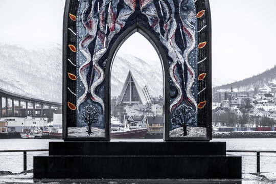 View of the Arctic Cathedral framed by a colorful mosaic archway with snowy mountains in the background in Tromso, Troms, Norway.