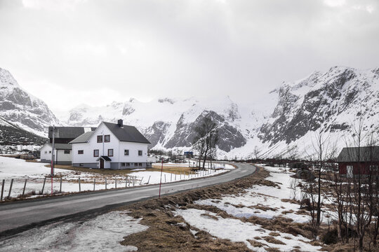 View of a traditional white house nestled in a snowy valley surrounded by majestic snow-capped mountains under an overcast sky in Kvaloya, Troms, Norway.