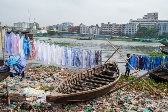 Dhaka Division, Bangladesh - 14 May 2026: View of the Buriganga River with a man hanging laundry on lines next to a wooden boat on a polluted bank.