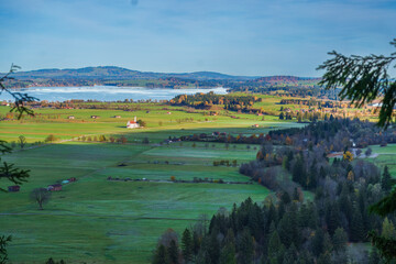 Fototapeta premium High angle view of St. Coloman church, green meadows, and Forggensee lake in autumn, seen from Neuschwanstein Castle, Bavaria, Germany.