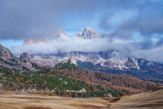 Beautiful autumn mountain landscape in Dolomites, Italy. Dramatic peaks partially covered by clouds above a colorful forest of golden larch and green pine trees on a sunny day.