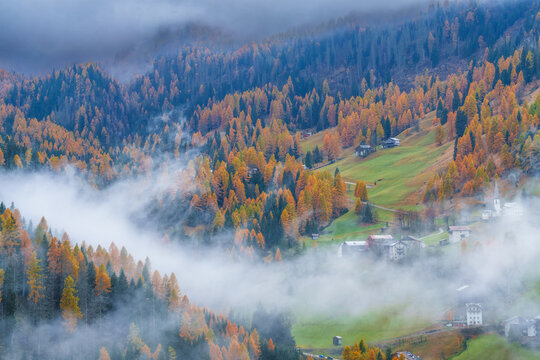 Beautiful autumn mountain landscape with a small village church in the Dolomites, Italy. Steep hillside covered with colorful golden larch forest partially obscured by thick fog and low clouds.