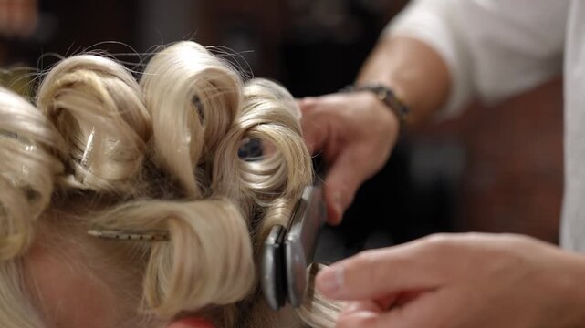 Close-up of a stylist using a hot iron to create beautiful, voluminous curls on a client's blonde hair in a modern beauty salon