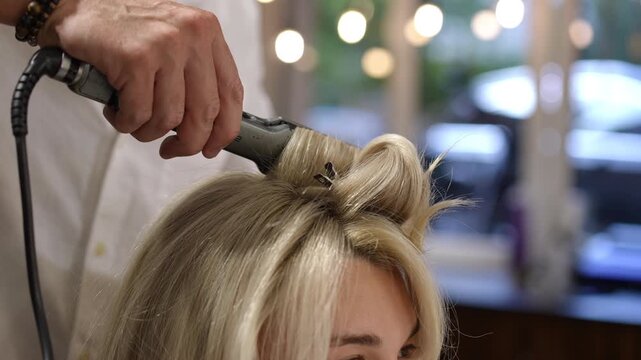 Close-up of a professional male hairdresser curling a blonde client's hair with a hot iron tong, creating a beautiful and stylish hairdo