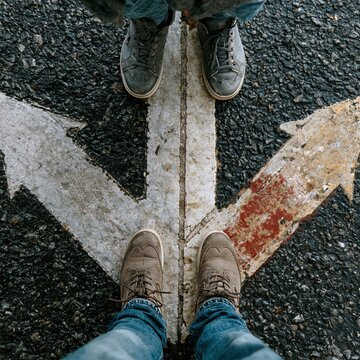 Close-up on the central split of a three-way arrow. Only the feet and legs of a figure are visible at the fork. Dramatic perspective