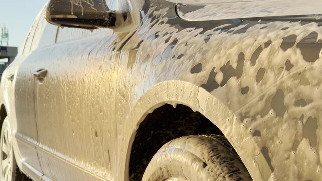 A modern grey car is covered in thick white foam during a wash. The soap drips down the vehicle, creating a satisfying and refreshing feeling of cleanliness.