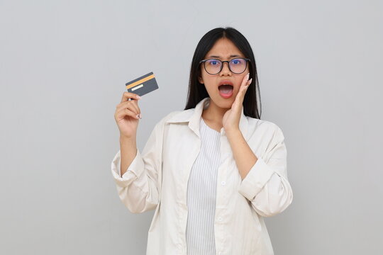 Photo of an impressed young Asian woman, hand on cheek, holding debit/credit card, surprised expression, screaming happily while looking at camera, isolated on white background.